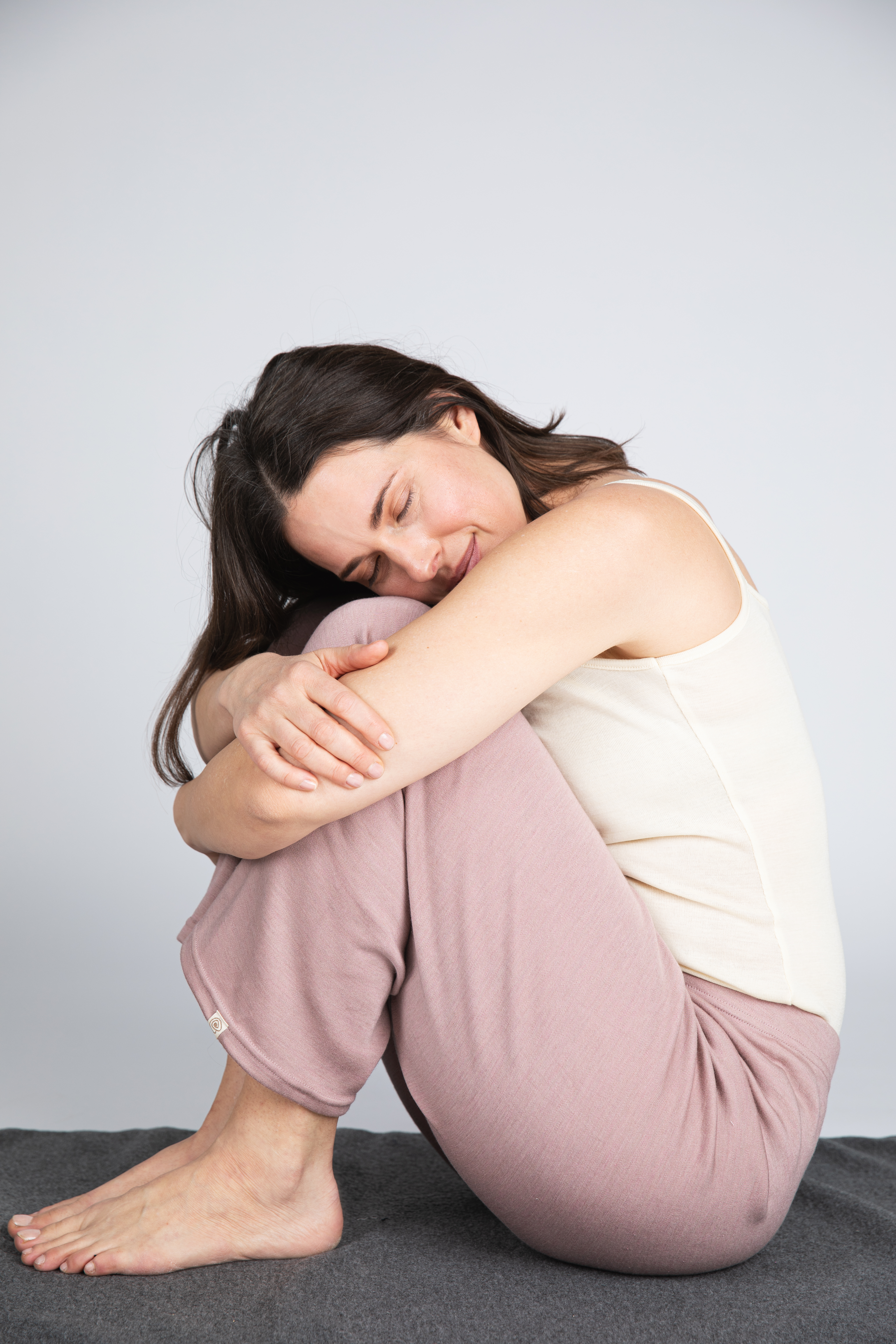  Frau trägt Woll-Seide-Interlock Loungehose in Dusty Rose von Engel, fühlt sich rundum wohl, sitzt barfuß und umarmt ihre Knie vor hellem Hintergrund.
